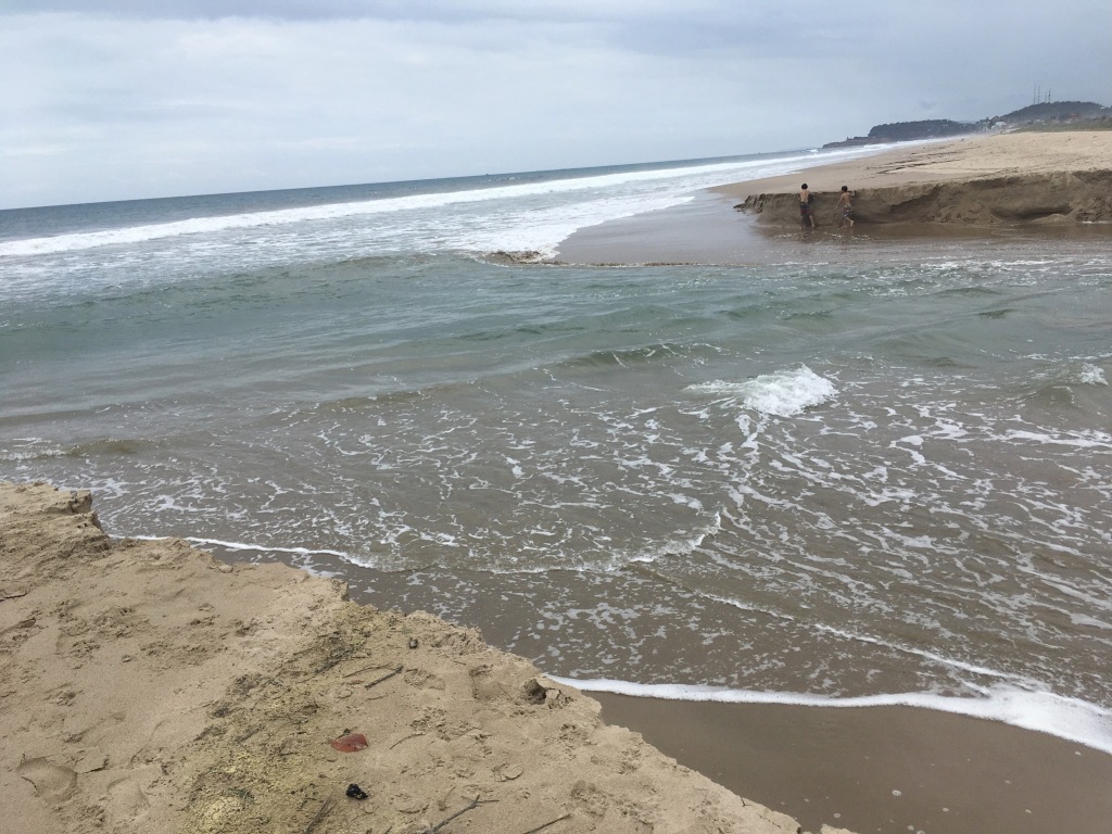 Photo of water flowing out to the ocean, carving out a widening breach in the sand.