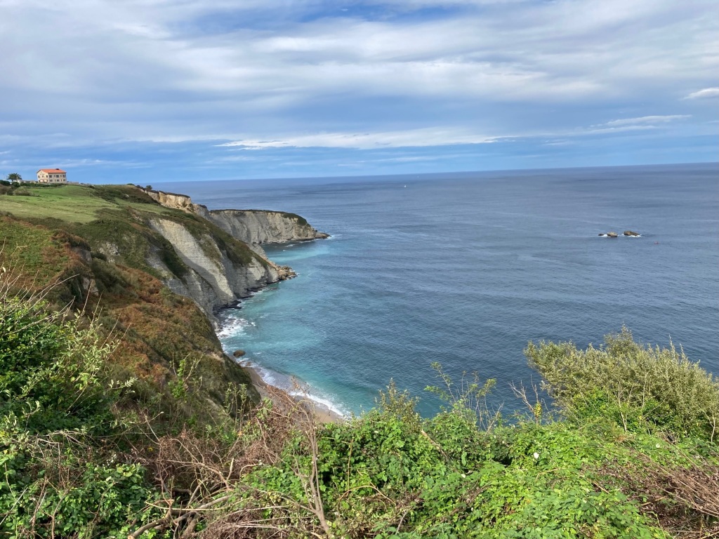 photo of gray cliffs rising out of teal-blue ocean with green fields on top and a stone inn on the point in the distance.