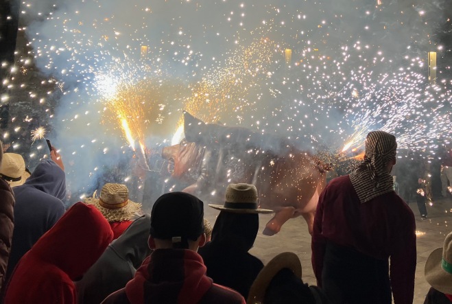 night photo of sprays of sparks shooting out of a large pig model surrounded by people in straw hats and bandanas
