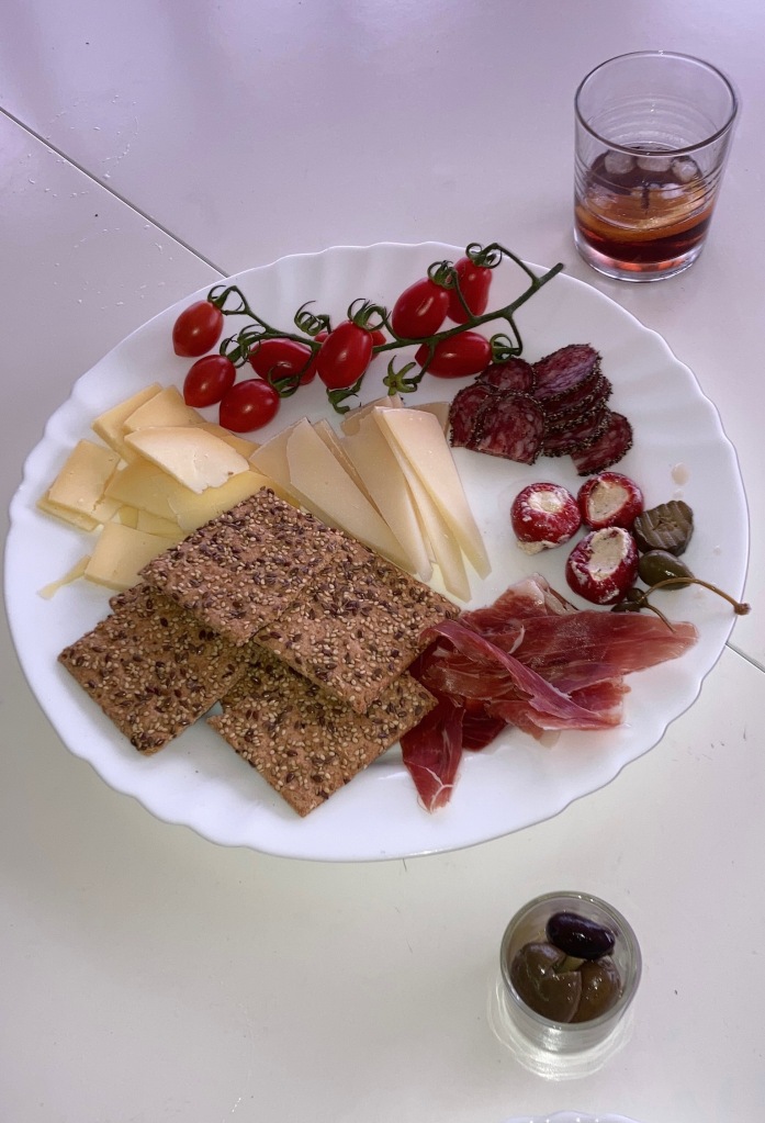 photo of a circular white plate with brown speckled crackers, an assortment of sliced white cheeses, red cherry tomatoes on a vine, slices of Iberian ham, olives, and small red peppers stuffed with white goat cheese, sitting on a white tabletop and flanked by two glasses of red vermouth.