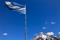 photo of a blue and white flag of Argentina blowing in the wind at the top of a flagpole with a blue sky and snow-capped volcano in the background