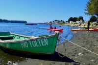 photo of a green wooden rowboat with RIO TOLTEN in white letters, resting on a dark sand beach. In the background is a wide blue river, a red kayak carrying two people lifting their paddles and an orange kayak on the beach with a person cheering next to it.
