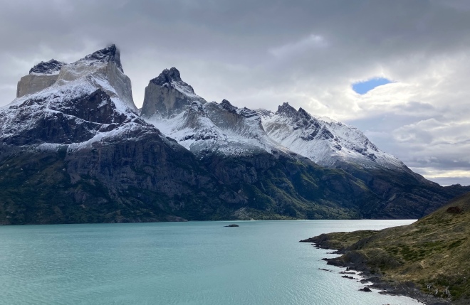 Photo of jagged brown mountain spires dusted with white snow above black cliffs and a glistening teal-green lake, with gray clouds and one small spot of blue sky above.