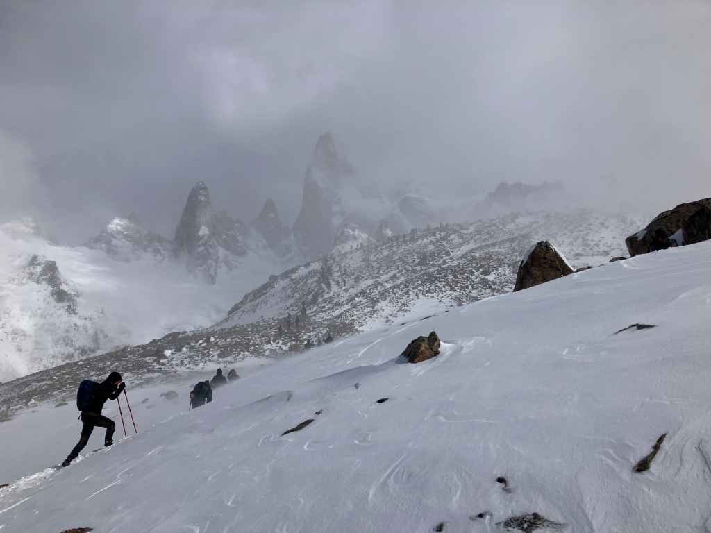 photo of a snowy mountain top with a struggling hiker in the foreground and cloudy rock spires in the background