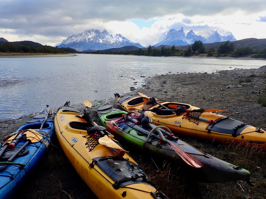 photo of four kayaks in blue, yellow and green, resting on gravel by the side of a gray river, with snow-capped mountains behind