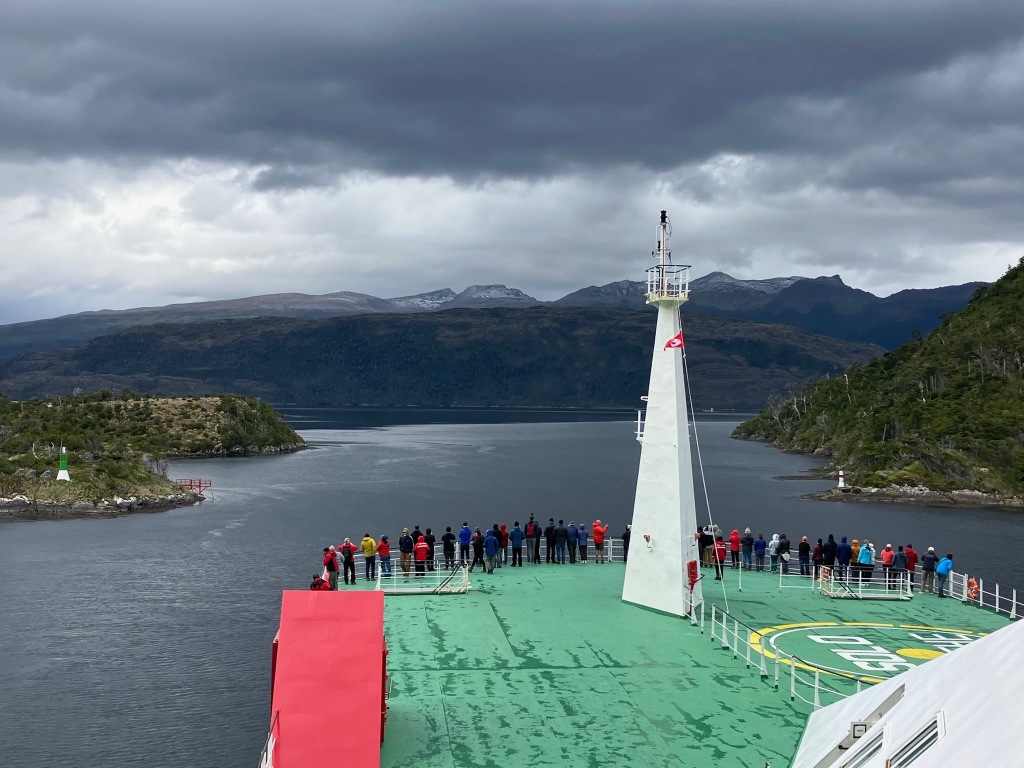 photo of the green and red deck of a wide ship with a white tower and a crowd of people on its bow, passing through a narrow channel with gray water, green hills on both sides and in the distance, and dark clouds above