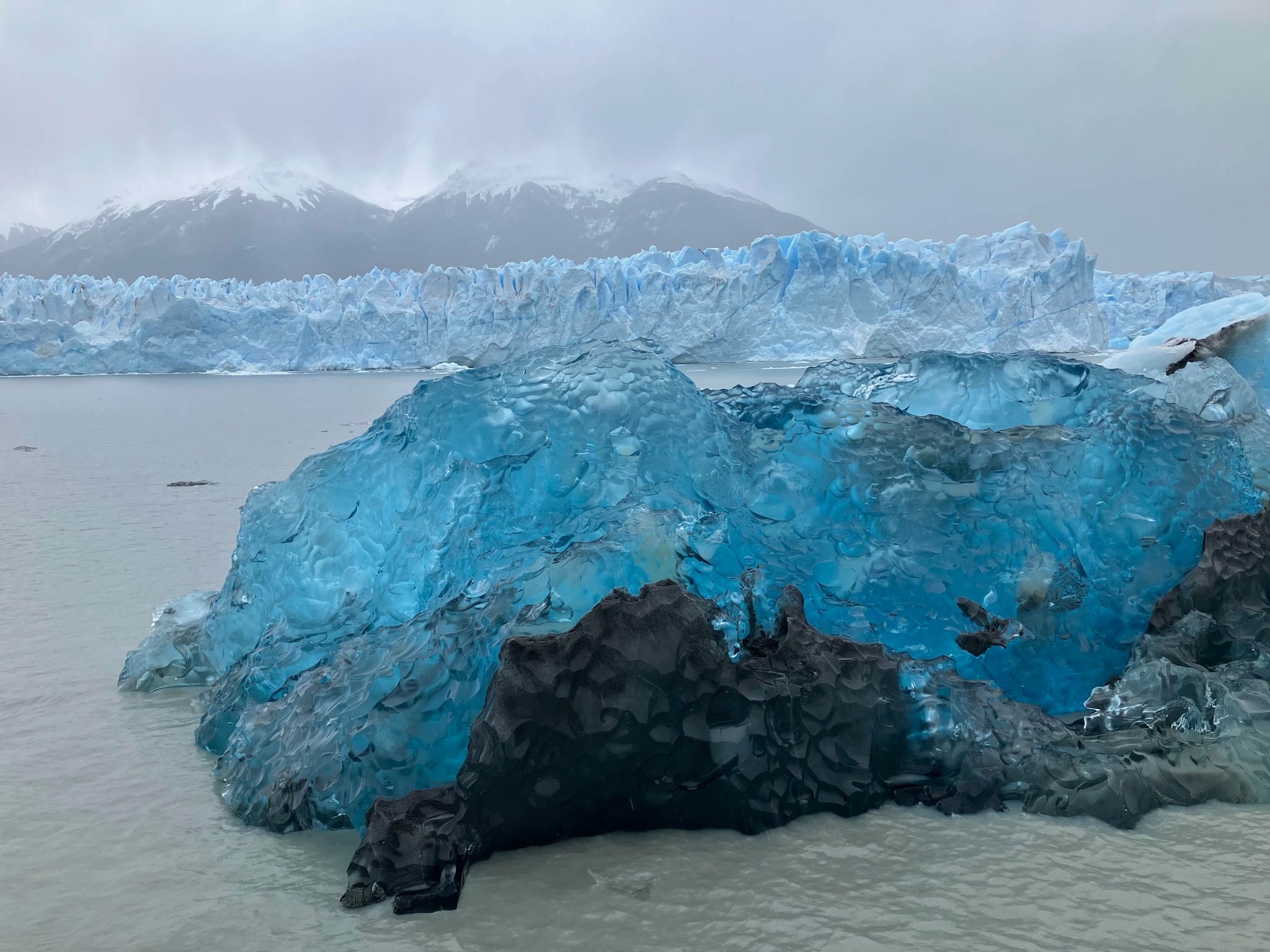 photo of a blue crystalline iceberg with a white glacier and mountains behind