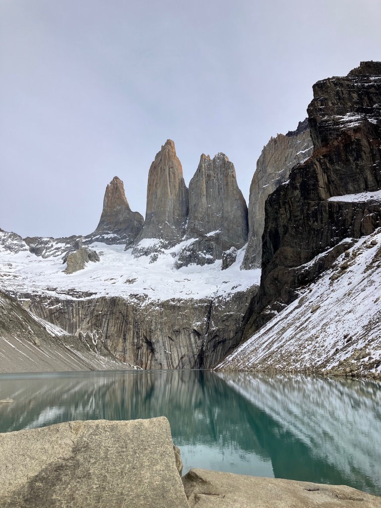 photo of three gray granite spires rising above a reflective green pond with gray rocks in the foreground and snow between